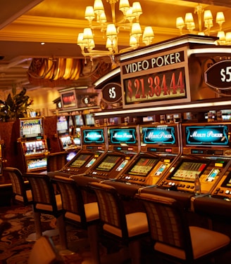 A casino floor featuring rows of brightly lit video poker machines. The area is illuminated by elegant chandeliers, with a display board showing a large sum of money, suggesting a possible jackpot. The rich decor includes luxurious carpets and comfortable seating, adding to the opulent atmosphere.