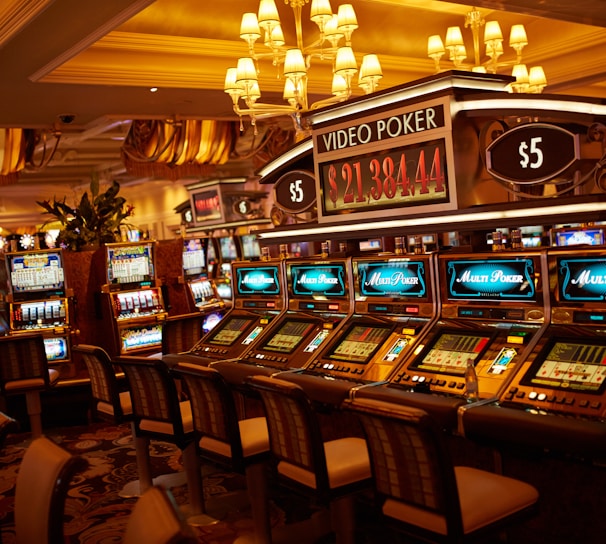 A casino floor featuring rows of brightly lit video poker machines. The area is illuminated by elegant chandeliers, with a display board showing a large sum of money, suggesting a possible jackpot. The rich decor includes luxurious carpets and comfortable seating, adding to the opulent atmosphere.